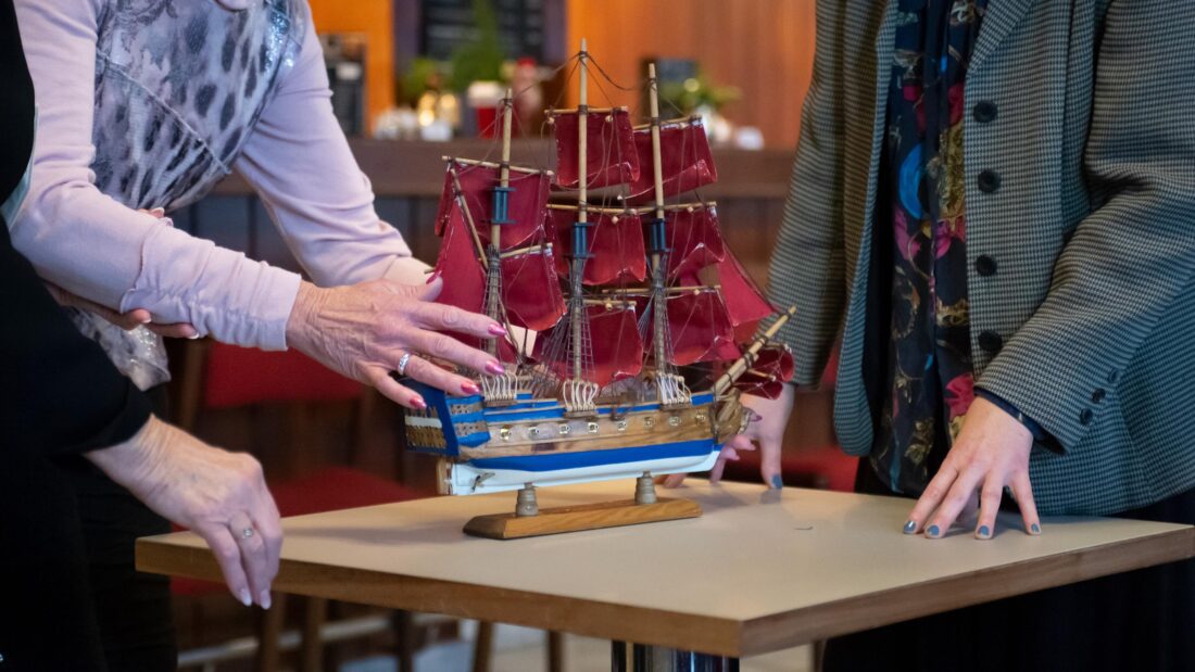 Auf dem Foto ist ein Theaterrequisit zu sehen: Ein Schiff, welches auf einem Holztisch steht und zwei Personen, die daneben stehen und erklären.