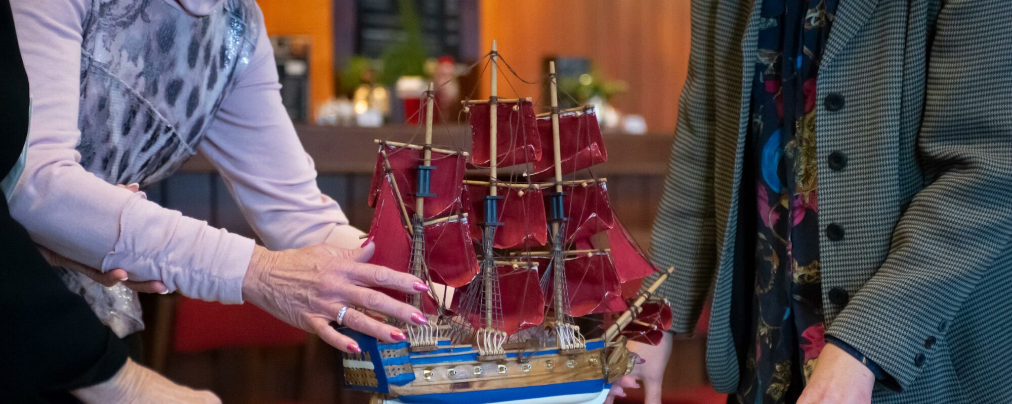 Auf dem Foto ist ein Theaterrequisit zu sehen: Ein Schiff, welches auf einem Holztisch steht und zwei Personen, die daneben stehen und erklären.